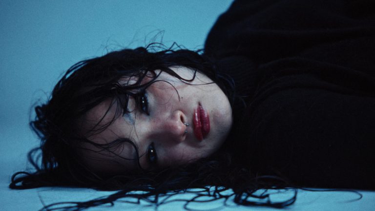 Woman with wet dark hair lies on a pale blue floor, looking into the lens, in the fashion film BLUE