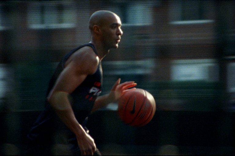 Basketball player dribbling past blurred housing blocks on a fast pan, in E x H – Chasing the Shot for Huel