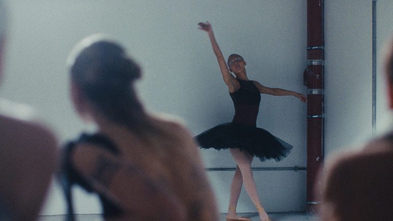 Ballerina in a black tutu dances against a white wall as seated dancers watch, from the fashion film Révérence